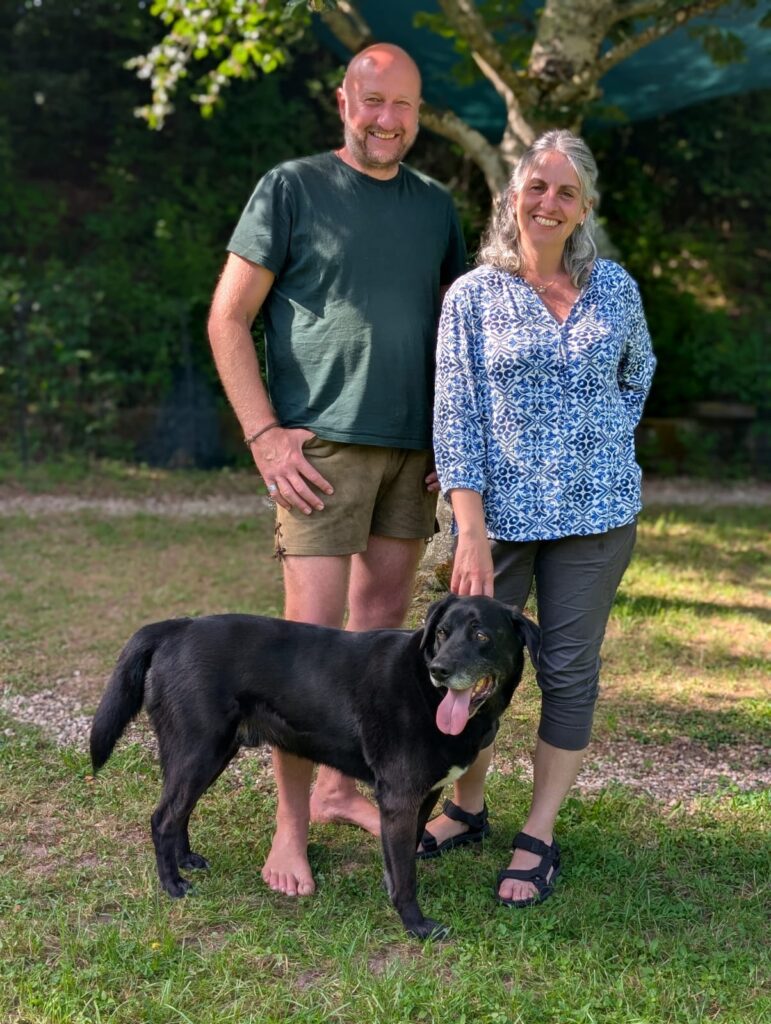 Smiling man and woman outdoors with a black dog standing on a grassy lawn.