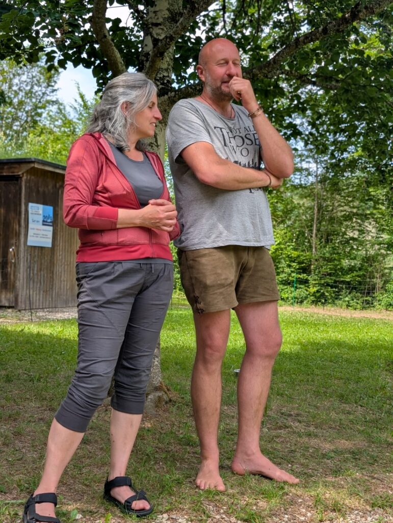 Two adults standing outdoors under a tree, chatting with arms crossed, a wooden shed in the background.