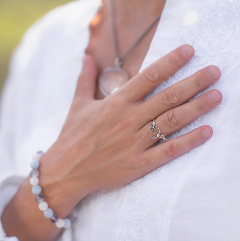 Close-up of a hand resting on the chest, wearing two silver rings and a beaded bracelet against a white blouse with a pendant visible