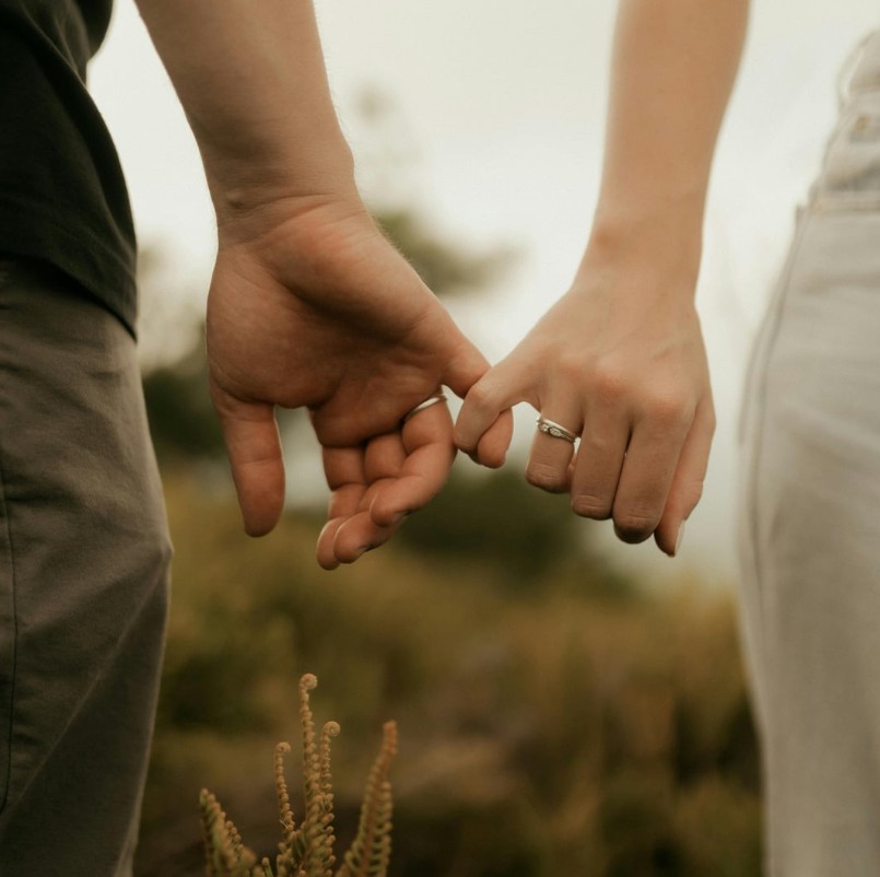 Two people holding hands outdoors; one hand has a ring on the ring finger with a blurred natural background in view.